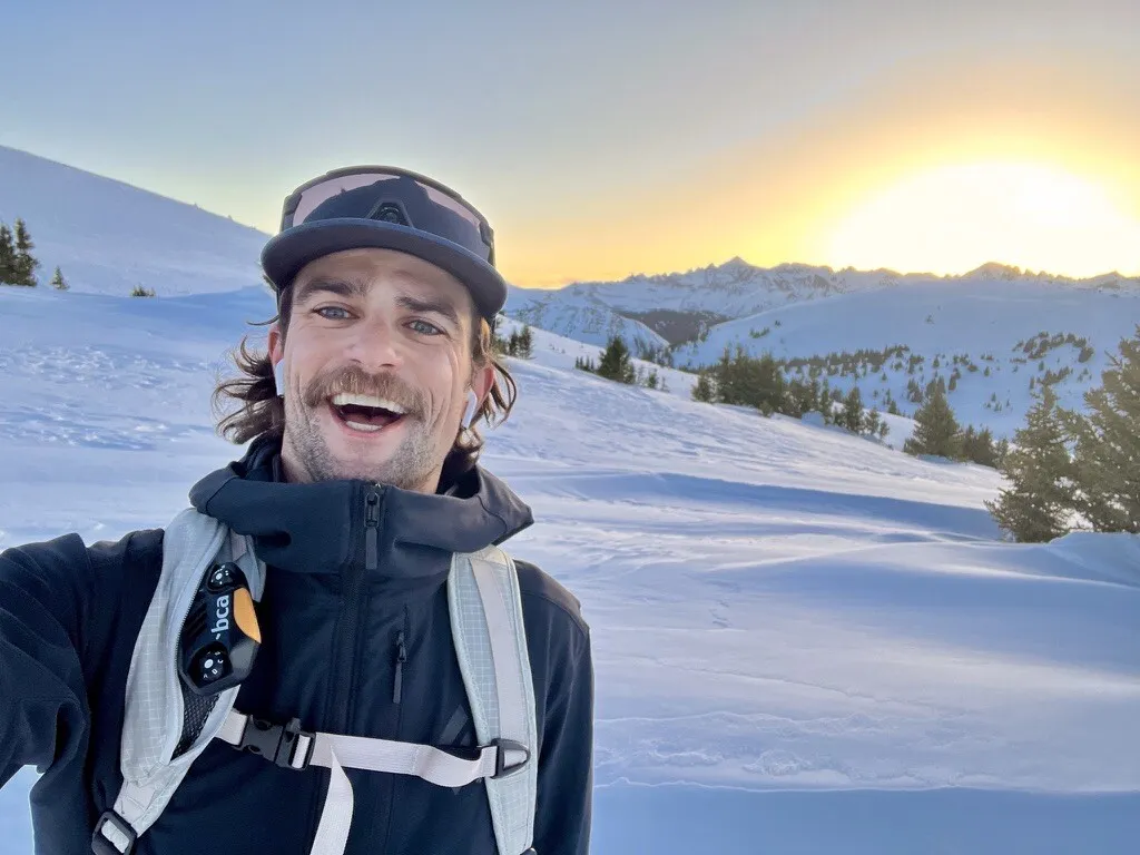Fletcher Keeley grinning on a ski tour at sunset, snowy peaks behind